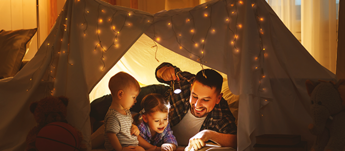 dad reading to his little children with a flashlight while laying in a tent in the family room